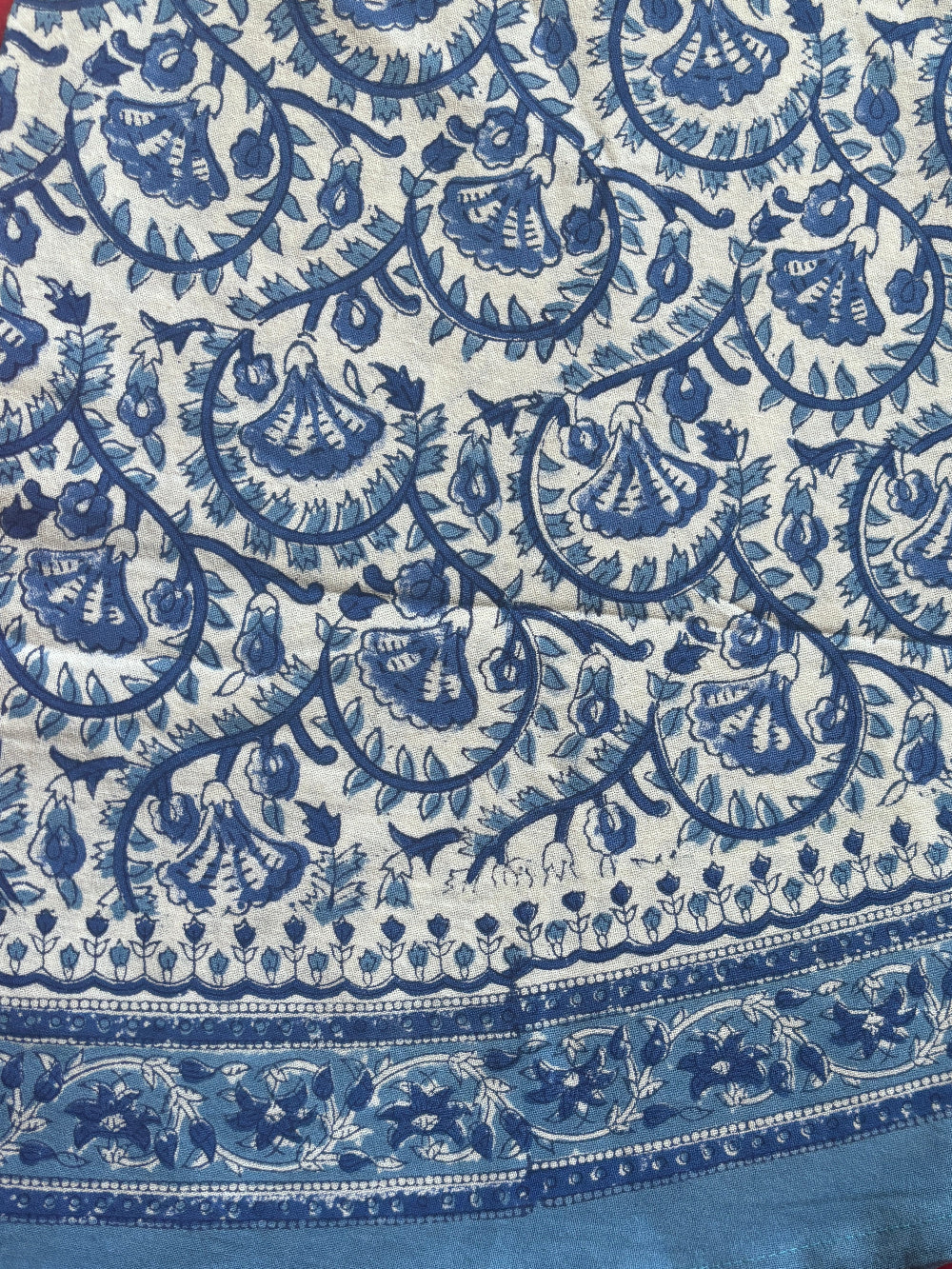 A close-up of a blue and white printed fabric with an intricate floral and paisley pattern, featuring a solid blue border, laid over a red patterned textile.