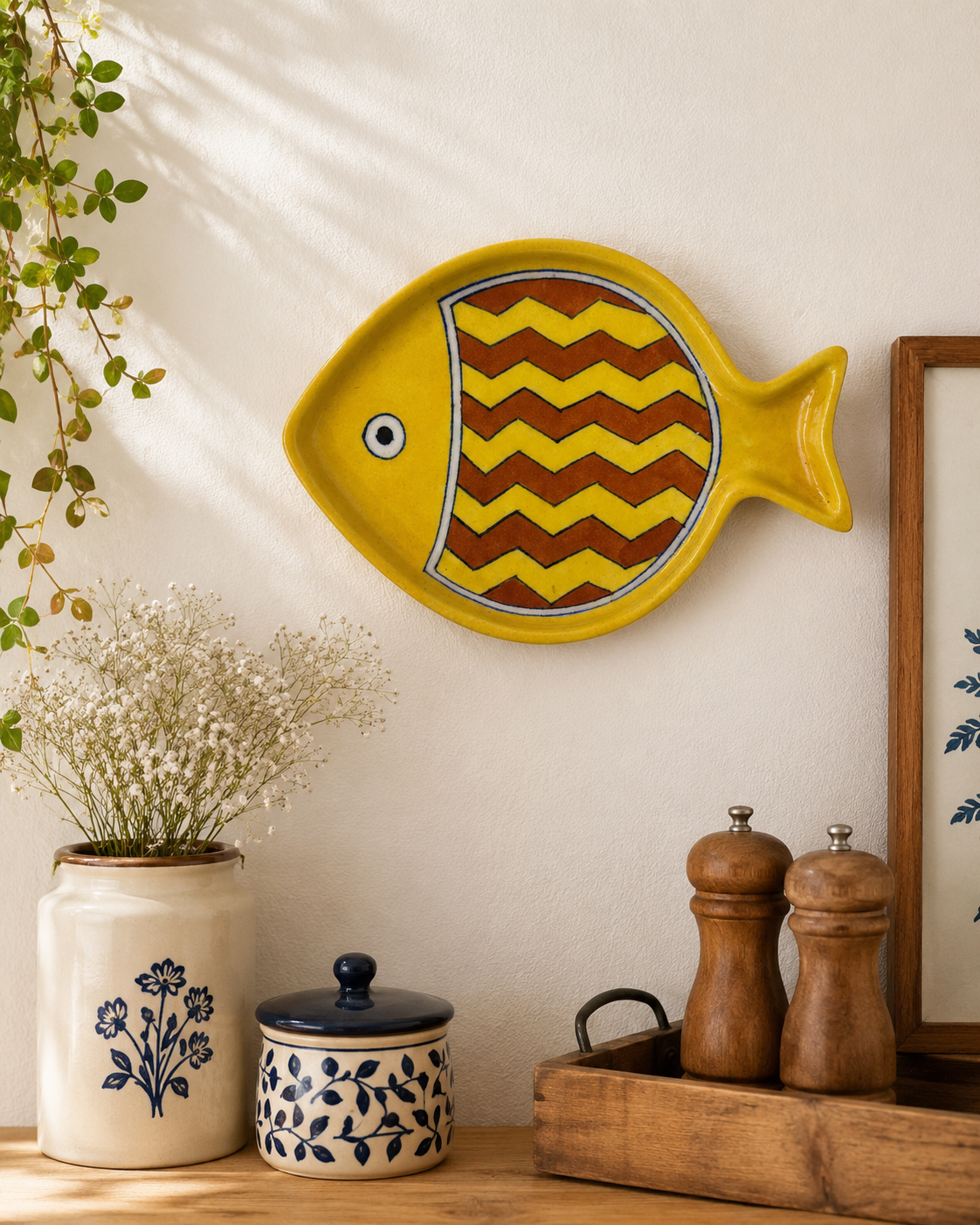 Yellow fish-shaped decorative plate on a wall with a plant and wooden tray below.