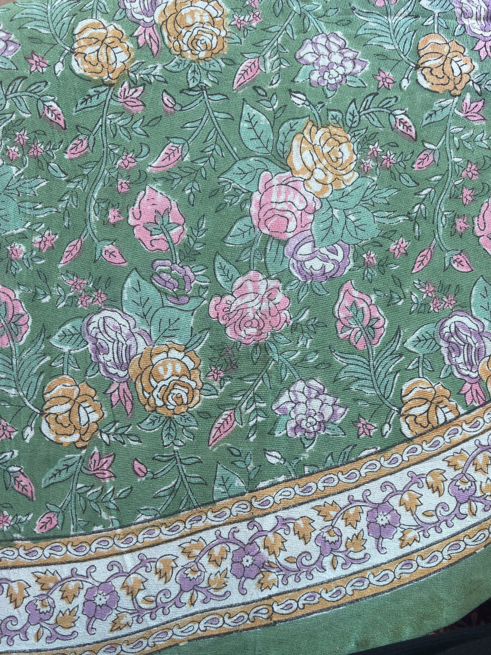 A close-up of a green fabric with a colorful floral print and a decorative border. The pattern features pink, yellow, and purple flowers with green leaves on a sage green background, and the border has a white base with gold and purple floral motifs.